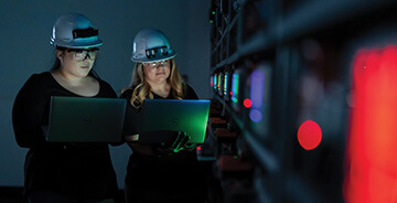 Two people wearing safety helmets working on laptops in a dimly lit server room with colorful indicator lights on the equipment.